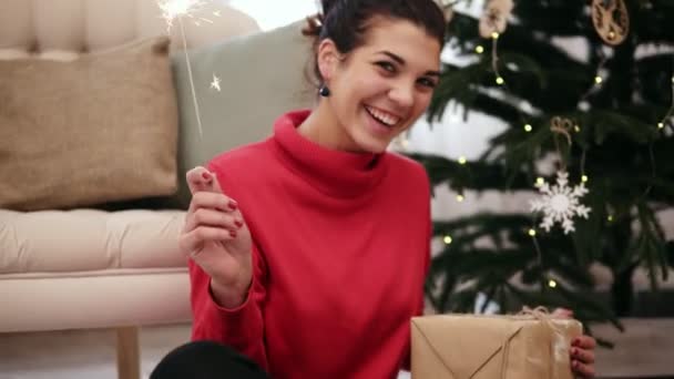 Feliz joven atractiva sentada en el suelo junto al árbol de Navidad celebrando la Navidad con bengalas y mirando a la cámara — Vídeos de Stock