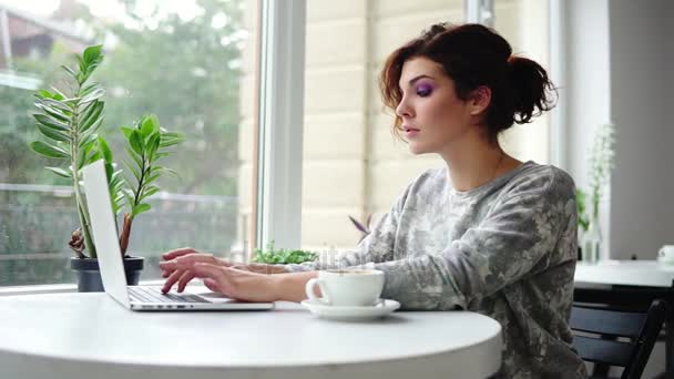 Jeune belle femme assise près de la fenêtre dans un café et travaillant sur un ordinateur portable. Fille surfant sur Internet et tapant. Tasse blanche avec café sur la table — Video