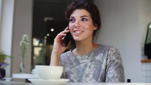 Женщина со смартфона разговаривает по телефону, сидя в кафе с чашкой кофе на столе. Она улыбается и смеется в кафе. Красивая молодая женщина с непринужденной беседой по мобильному телефону — стоковое видео
