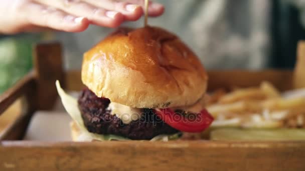 Sabrosa hamburguesa con papas fritas en una bandeja de madera. Las manos femeninas lo tocan para empezar a comer. Disparo en cámara lenta — Vídeo de stock