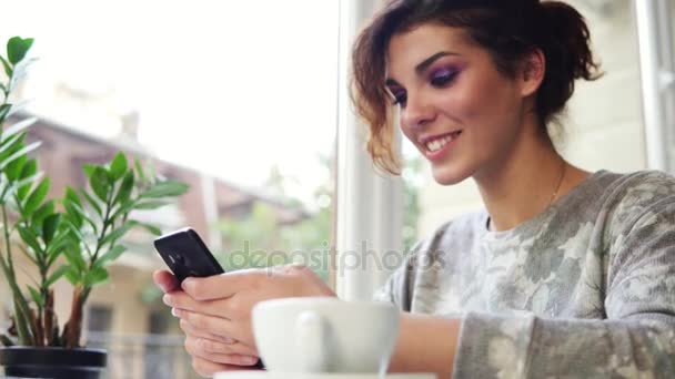 Mujer atractiva joven de compras en línea utilizando su teléfono móvil en la cafetería y sonriendo. Mujer usando la aplicación en el teléfono inteligente en la cafetería beber café — Vídeos de Stock