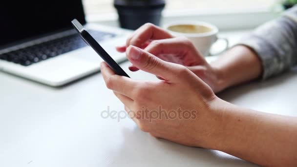 Крупный план женских рук с помощью приложения на смартфоне, сидя в кафе с чашкой кофе и ноутбуком на столе — стоковое видео