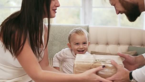 Joven madre vestida de blanco recibiendo regalo de Navidad de su marido y besándolo. Rubia lindo niño mirando a sus padres y sonriendo. Fiesta de Navidad familiar en casa — Vídeo de stock