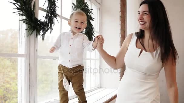 Ung leende mamma i vit klänning håller sin sons hand medan han går på fönsterbrädan dekorerad med Christmas Krans. Lycklig familj kyssar — Stockvideo