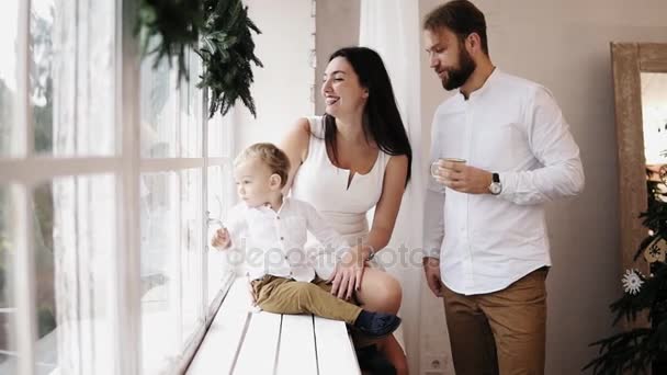 Jonge mooie familie zitten samen bij het raam versierd met de kroon van Kerstmis. Liefhebbende ouders elkaar zoenen in de ochtend na Kerstmis — Stockvideo