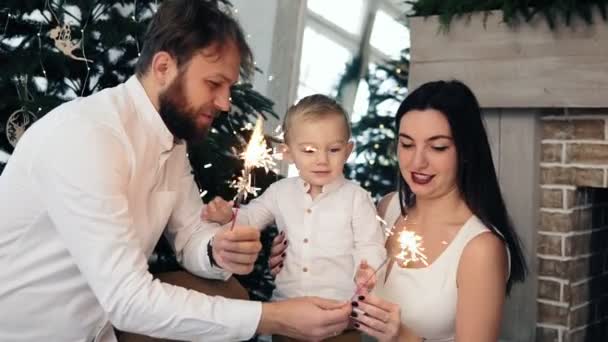 Familia feliz y fuego de Bengala en casa junto al árbol de Navidad. Madre y padre sosteniendo chispeante, mientras que su lindo hijo está mirando la luz. Preciosa familia celebrando la Navidad en casa — Vídeos de Stock