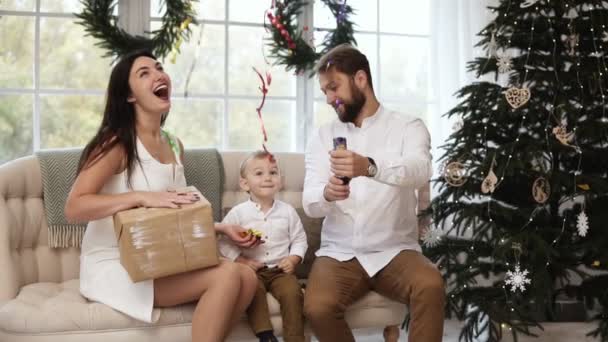 若い母親が彼女の手にプレゼントを持っている若い父親は花火、キラキラと宙を舞う紙吹雪を爆破しながら。陽気な家族のクリスマス ツリーがかわいい幼児 — ストック動画