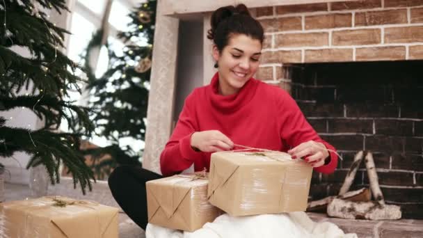 Красивая улыбающаяся женщина заворачивает рождественские подарки сидя у елки и камина дома, а затем кладет их под елку. Рождественская подготовка — стоковое видео