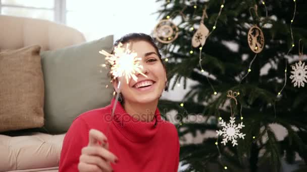 Χαμογελώντας φιλικό ελκυστική νεαρή γυναίκα που κάθεται στο πάτωμα από το χριστουγεννιάτικο δέντρο που γιορτάζει τα Χριστούγεννα με βεγγαλικά και ψάχνει στην κάμερα — Αρχείο Βίντεο