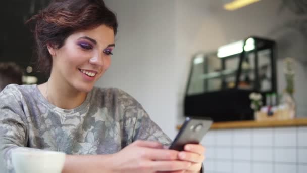 カフェで彼女の携帯電話を使って美しい若い女性の側面図です。笑みを浮かべて。女性が携帯電話でコーヒー カフェを飲むでスマート フォン上のアプリ、テキスト メッセージを使用して — ストック動画