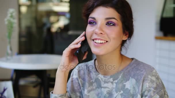 Смартфон жінка розмовляє по телефону, сидячи в кафе. Вона посміхається. Красива молода жінка має випадкову розмову на мобільному телефоні — стокове відео