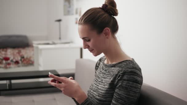 Retrato de uma jovem feliz sentada no sofá em casa olhando para o telefone e digitando uma mensagem — Vídeo de Stock