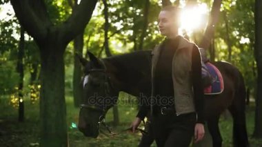 Çekici genç kadın jokey beyaz nokta ile kahverengi at ile ormanda alnına sonbahar deri kayışı eyer tutan güneşli bir günde sırasında yürümekten Slowmotion vurdu