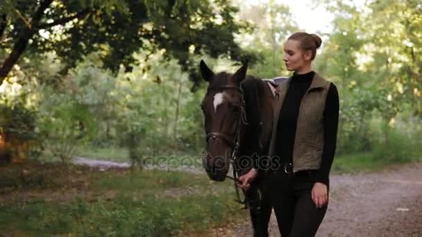 Jovem mulher bonita está acariciando um cavalo marrom deslumbrante com mancha branca na testa enquanto caminham juntos na floresta durante o dia ensolarado no outono — Vídeo de Stock
