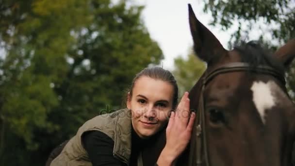 Giovane bella donna accarezzando cavallo marrone mentre seduto in sella e mettendo la testa sulla testa dei cavalli guardando nella fotocamera — Video Stock