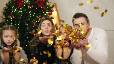Dört altın glitter konfeti evde ağır çekimde Noel partisi sırasında üfleme mutlu aile. Arka planda bir şekilde dekore edilmiş Noel ağacı. Slowmotion atış