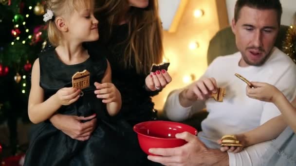 Lycklig familj av fyra sitter vid julgranen och äta choklad kakor. Söt mamma, Pappa och två döttrar firar jul tillsammans. Slowmotion skott — Stockvideo