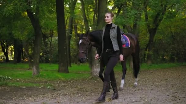 Joven jinete de caballo feliz está caminando con caballo marrón con mancha blanca en la frente en el parque durante el día soleado sosteniendo correa de cuero de silla de montar en su mano. Verdadera amistad — Vídeos de Stock