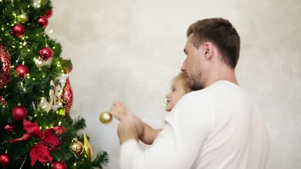 Joven padre sosteniendo a su linda hijita en sus brazos ayudándola a colgar una bola de oro en un árbol de Navidad. Decorar el árbol de Navidad juntos en casa. Movimiento lento — Vídeos de Stock