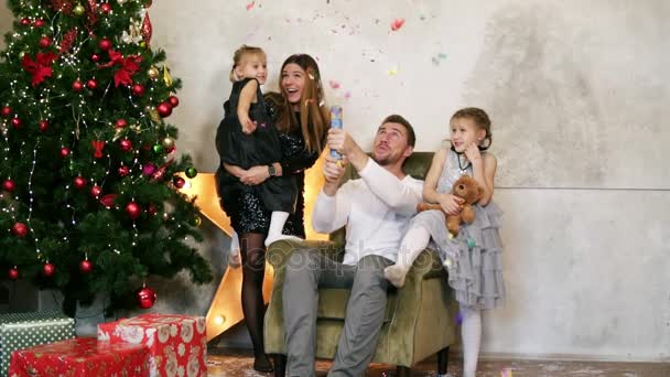 父、母と 2 人の娘が一緒にクリスマスを祝ってします。4 つ飾られたクリスマス ツリーのそばに座って petards を持つ楽しみのかわいい家族 — ストック動画