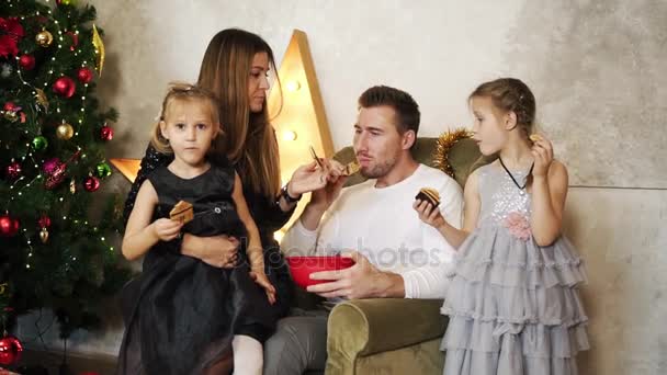 Счастливая семья из четырех человек сидит у елки и ест шоколадное печенье. Милая мать, отец и две дочери празднуют Рождество вместе. Слоумоушн — стоковое видео