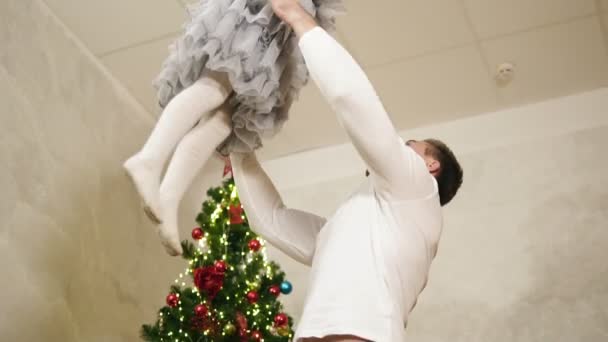 Νεαρός πατέρας ρίχνει την αξιολάτρευτη κόρη του στον αέρα στο σπίτι με το στολισμένο χριστουγεννιάτικο δέντρο στο παρασκήνιο. Είναι χαμογελαστός και χορό όταν πίσω στο πάτωμα — Αρχείο Βίντεο