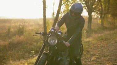 Kask ve deri ceketli genç adam bisikletini için gelir ve sonbaharda güneşli bir günde kenarında dururken ait değilsin. Slowmotion atış