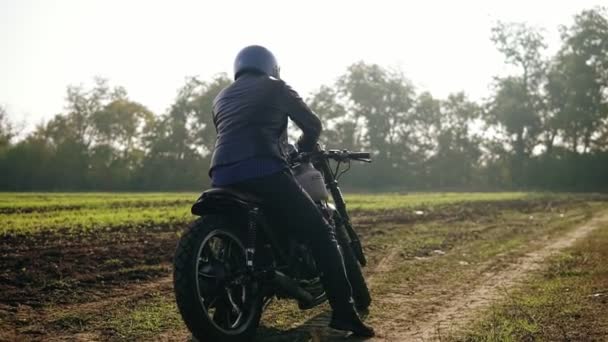 Vista trasera de un hombre con casco y chaqueta de cuero acercándose a su bicicleta y arrancando el motor mientras está parado en el campo abierto en un día soleado. Disparo en cámara lenta — Vídeo de stock