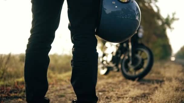 Visão traseira de um homem irreconhecível em jeans pretos e jaqueta de couro segurando seu capacete na mão enquanto estava de pé em sua bicicleta legal em um dia ensolarado no outono na floresta — Vídeo de Stock