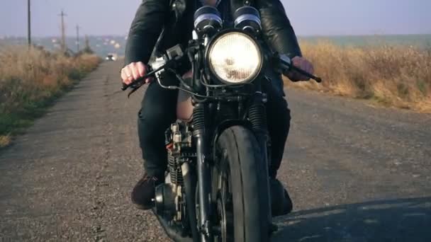 Front view of a unrecognizable man in black leather jacket riding motorcycle on a country road holding a handlebar. Big headlight turned on — Stock Video