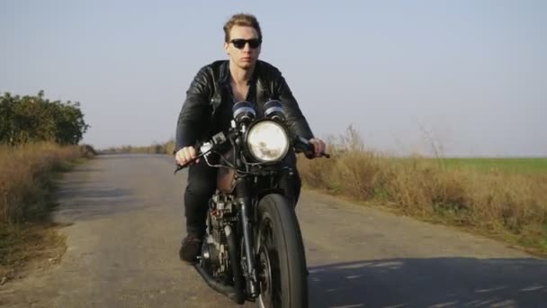 Portrait of a stylish cool young man in sunglasses and leather jacket riding motorcycle on a road on a sunny day — Stock Video