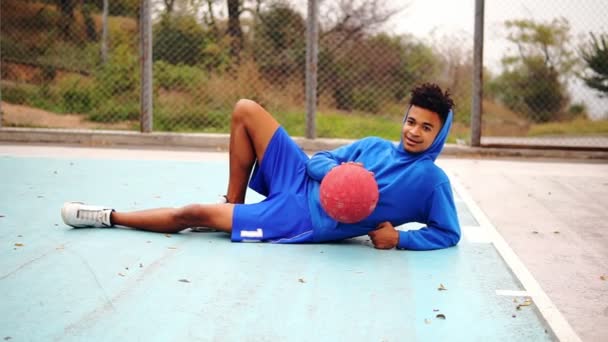 Joven afroamericano tendido en el suelo del campo de juego y jugando con la pelota de baloncesto. Disparo en cámara lenta — Vídeos de Stock