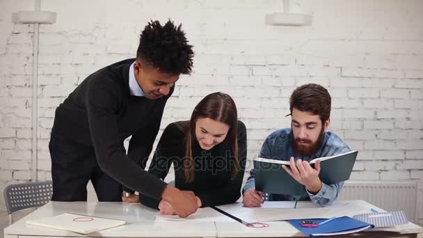 Mujer joven está dibujando un plan con lápiz sentado a la mesa. Su colegial africano le ayuda a terminar su trabajo mientras está de pie cerca de ella. Feliz grupo diverso de colegios que trabajan en el proyecto — Vídeos de Stock