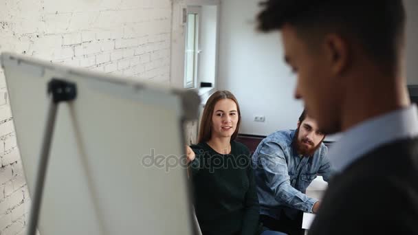 Vista posterior del oficinista afroamericano dando una presentación para sus colegas en la oficina en el rotafolio. Joven trabajador preguntando en el sofá y él está respondiendo — Vídeos de Stock