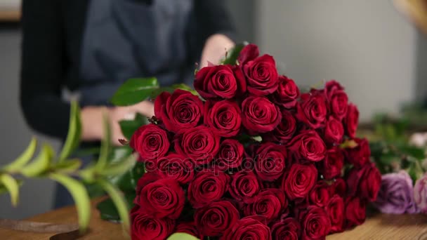 Um bando perfeito de rosas vermelhas na mesa. Vista de perto das mãos do florista feminino que arranja buquê moderno usando belas rosas vermelhas na loja de flores. Tiro em câmara lenta — Vídeo de Stock