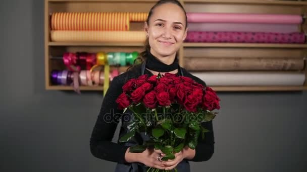 Ramo perfecto de rosas rojas de florista profesional: florista femenina atractiva joven sosteniendo un ramo de hermosas rosas rojas en la tienda de flores y mirando en cámara. Disparo en cámara lenta — Vídeos de Stock