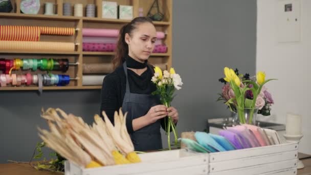 Молода жінка-флорист організовує сучасний букет у квітковому магазині, що стоїть за столом. Вона поєднує кремові троянди і жовті тюльпани — стокове відео