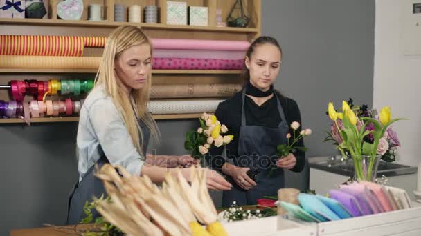 Aantrekkelijke blonde bloemist in schort staande met haar collega aan balie in floral winkel terwijl bos bloemen schikken — Stockvideo