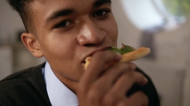 Vista da vicino del giovane afroamericano che gusta la pizza durante il pranzo al lavoro. Sta mordendo una fetta e guardando la telecamera al rallentatore — Video Stock