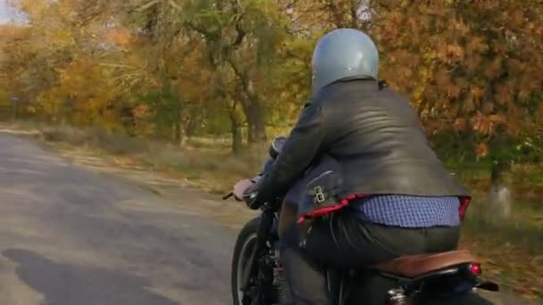 グレーのヘルメットと秋の晴れた日にアスファルトの道路でバイクに乗って革ジャケットと格子縞のシャツの男の背面します。茶色の木と黄色の葉道路周辺 — ストック動画