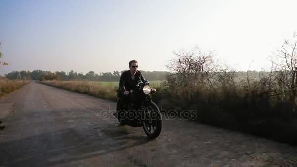 Elegante joven fresco en gafas de sol y chaqueta de cuero a caballo motocicleta en un camino de asfalto en un día soleado en otoño — Vídeos de Stock