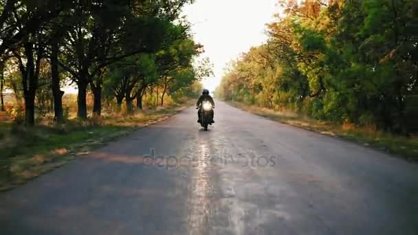 Vista frontal de hombre con estilo en casco negro y chaqueta de cuero a caballo motocicleta en una carretera de asfalto en un día soleado en otoño. Hermosos árboles con hojas amarillas y marrones alrededor de la carretera — Vídeos de Stock