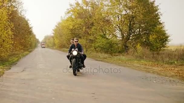 Tracking shot de pareja a caballo motocicleta en carretera forestal en otoño. Atractivo joven con gafas de sol conduciendo su helicóptero mientras su novia está sentada detrás — Vídeo de stock