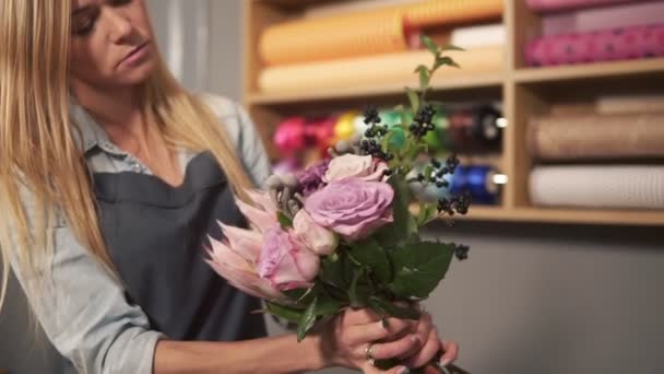 Jovem florista loira feminina organizando buquê moderno e olhando como as flores são combinadas. Bonito dono de loja de flores trabalhando em sua loja de flores fazendo buquê. Tiro em câmara lenta — Vídeo de Stock