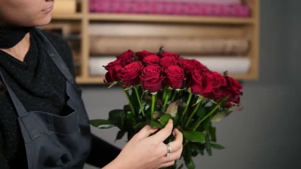 Concéntrate en el ramo de rosas rojas: florista femenina joven y atractiva que organiza un ramo de hermosas rosas rojas en la florería. Disparo en cámara lenta — Vídeo de stock