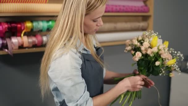Молодая блондинка-флористка завязывает кучу желтых тюльпанов и сливочных роз с лентой в руках — стоковое видео