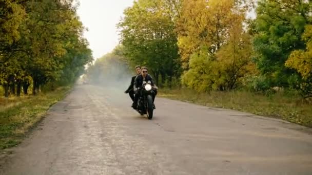 Hombre guapo en gafas de sol que monta con su novia en una motocicleta en el camino de asfalto en el bosque en otoño. Su novia tiene una granada de humo. Lento. — Vídeo de stock