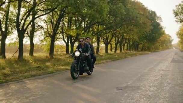 Seguimiento de tiro de joven pareja fresca a caballo motocicleta en carretera forestal en otoño. Atractivo joven con gafas de sol conduciendo su helicóptero mientras su novia está sentada detrás — Vídeos de Stock