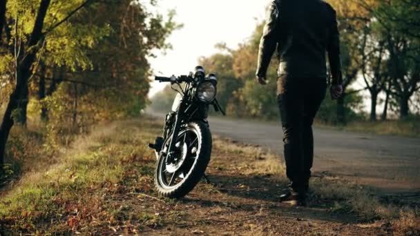 Uomo irriconoscibile in casco e giacca di pelle che arriva fino alla sua moto e avvia il motore mentre in piedi sul ciglio della strada in una giornata di sole in autunno — Video Stock