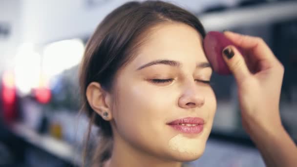 用海绵在年轻女子脸上应用液体基础的不可辨认 visagist。专业化妆沙龙。Slowmotion 射击 — 图库视频影像